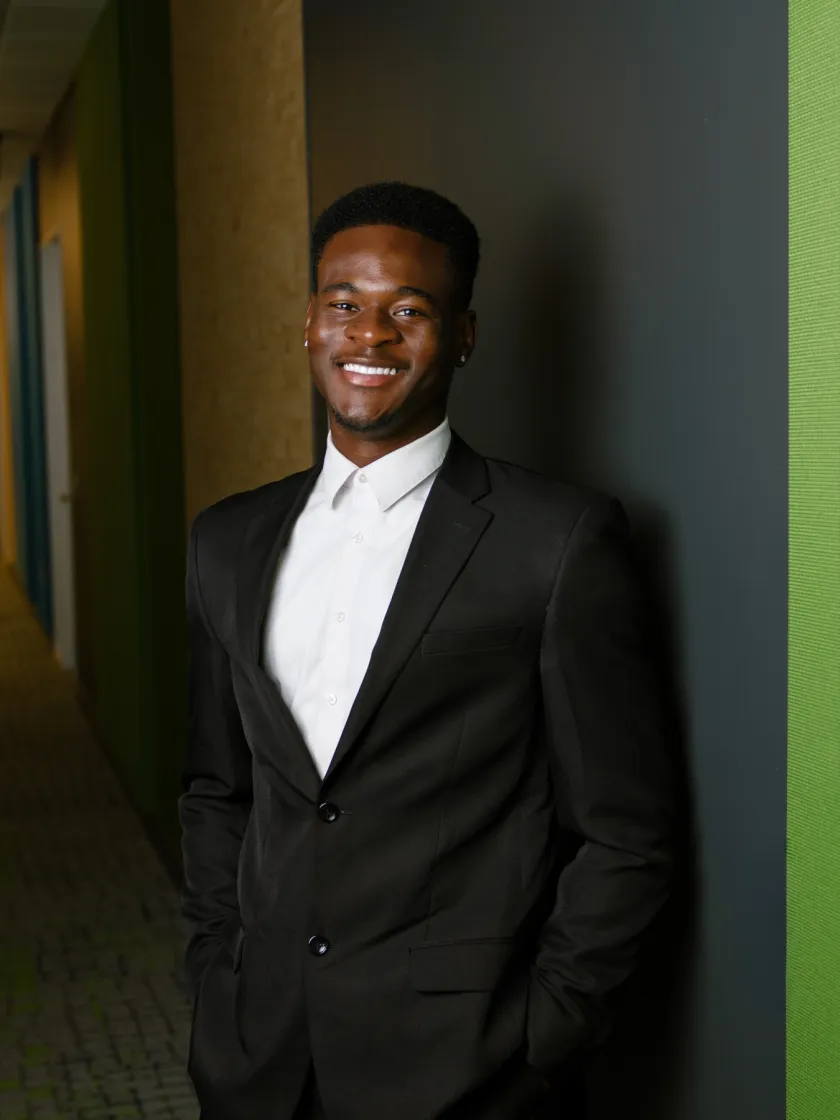 Daniel, smiling, in a dark suit and white shirt, leaning against a green wall